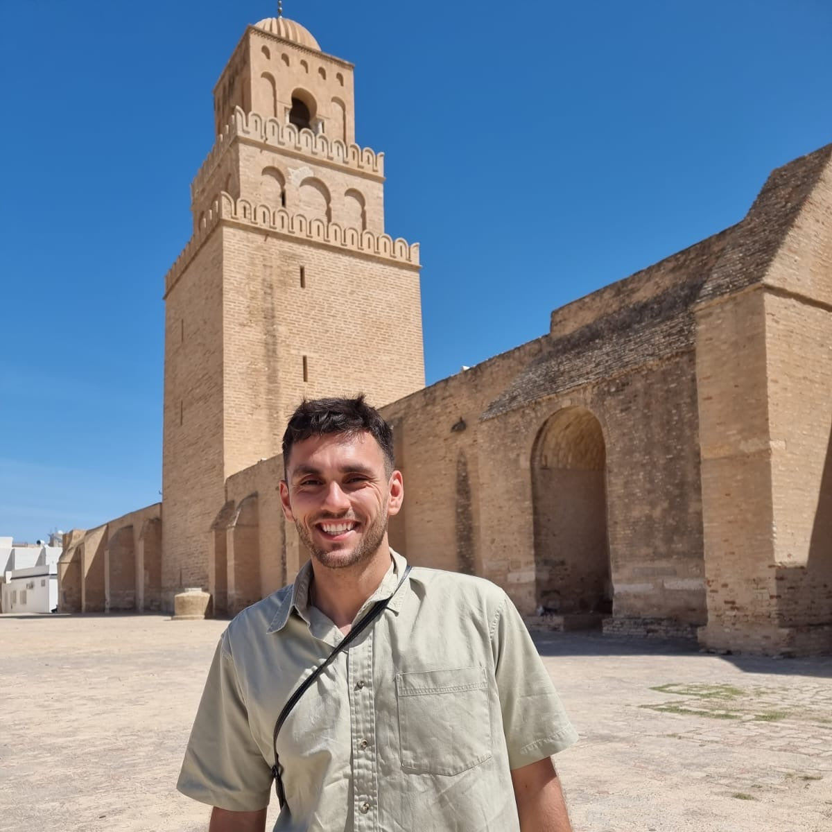 Man standing in front of a large brick building with a minaret under a clear blue sky.
